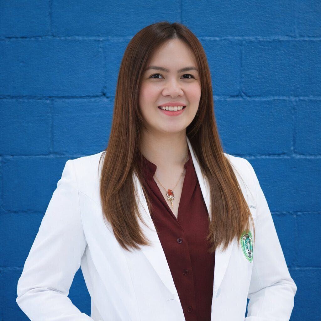 Professional woman in a white coat and burgundy blouse smiling, standing with hands on hips against a blue wall.